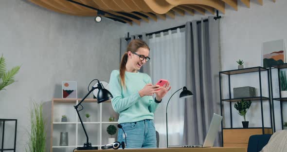 Girl in Casual Clothes Standing Near Table in Living-Room and Uses Her Mobile to Watch Funny Videos alt