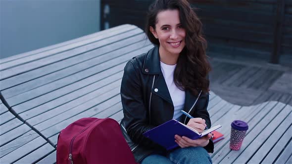 Female Student Writing in Notebook Sitting Outside on the Modern Wooden Bench alt