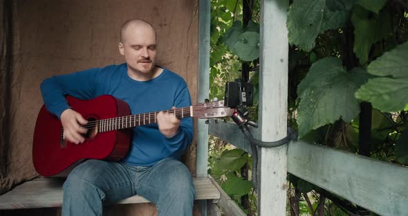 Man with Acoustic Guitar Sits on Porch of Village House and Hosts Online Podcast