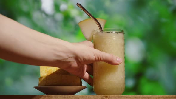 Female Hand Puts Glass of Melon Smoothie on Table Bar alt