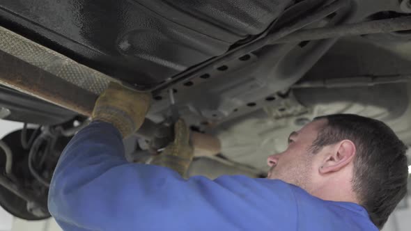 Experienced Auto Mechanic in a Blue Uniform Standing Under the Car with a Wrench and Tightening the alt