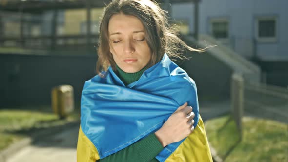 Sad Young Woman Wrapped in a Blue and Yellow Flag of Ukraine Stands with Her Eyes Closed alt