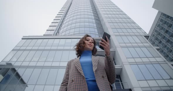 Young Woman in Jacket Stands at the Office Center and Gets Text Message in Her Phone Woman Smiles alt