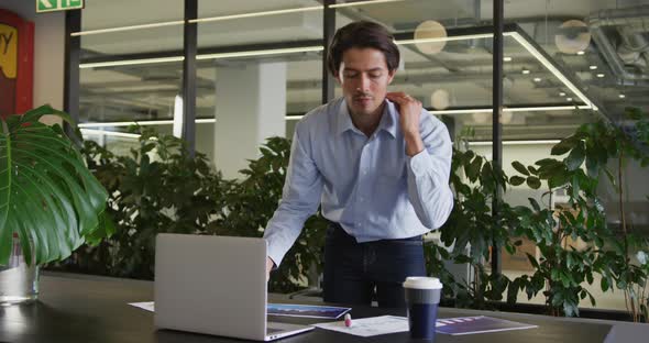 Caucasian businessman standing using laptop going through paperwork in modern office alt
