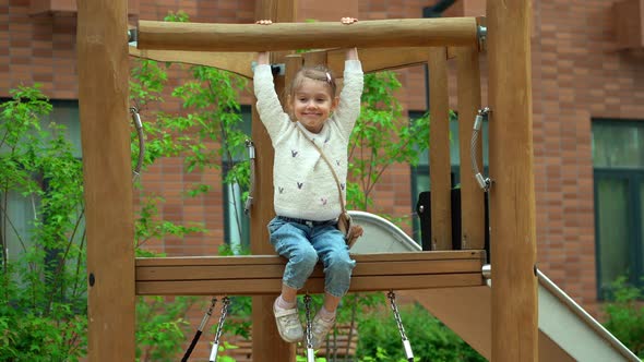 Little Girl Sits on Slide on Playground Looks at Camera Smiles and Laughs in Green Summer Park