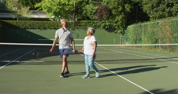 African american senior couple holding rackets talking to each other on the tennis court alt