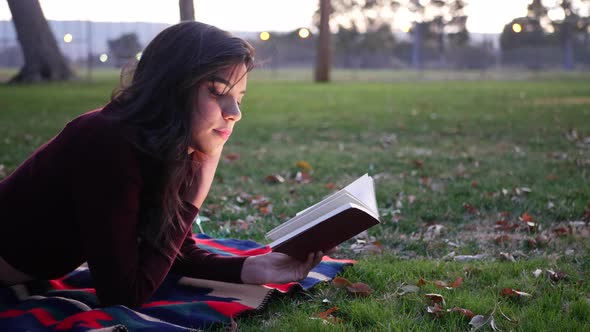 A young hispanic woman reading a book and relaxing in the park at sunset. alt