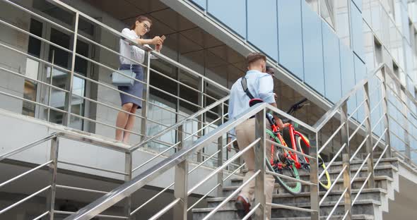 Caucasian Businessman Climbing Upstairs with Bicycle and Greeting Colleagues Outside Office Building alt