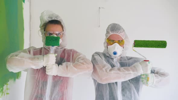 Couple in Protective Suits Poses with Paint Rollers in Room alt