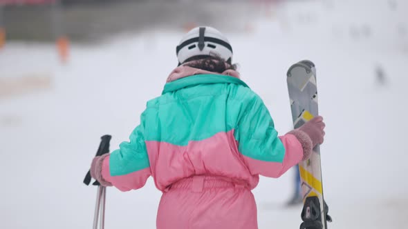 Back View of Slim Woman in Ski Suit and Helmet Walking in Slow Motion with Skis and Ski Poles alt