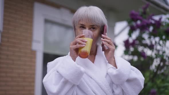 Confident Mature Woman Talking on Phone Drinking Orange Juice Standing on Backyard Porch alt