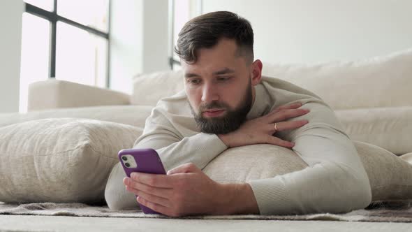 Bearded Young Man Uses a Smartphone Lying on the Floor of the House alt