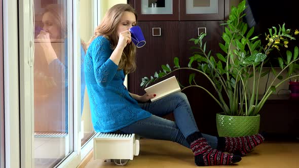 Lovely Woman Read Book and Drink Coffee Sitting on Warm Radiator