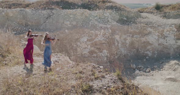 Far View of Two Elegant Musicians in Dresses Play Violin Among Cliff in Summer alt
