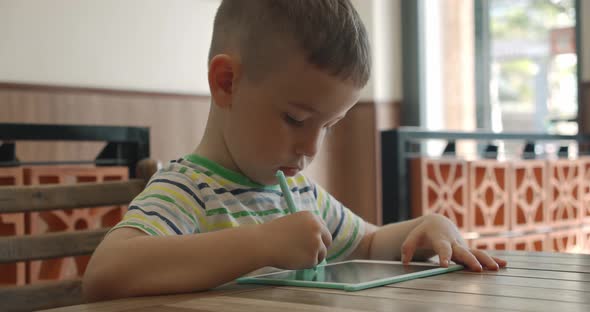 A Portrait of a Child Sits at a Table at Home and Draws on His Tablet with a Special Pen a alt