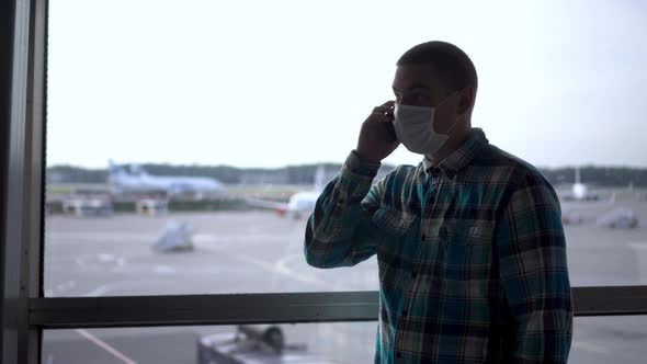 A Young Man in a Medical Mask Talks on the Phone Against the Background of a Window at the Airport alt