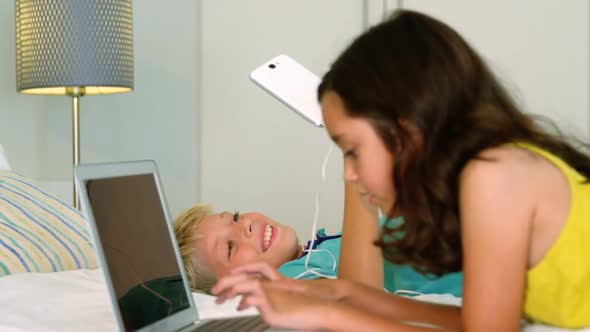 Siblings sitting back to back and using digital tablet, laptop in bedroom alt