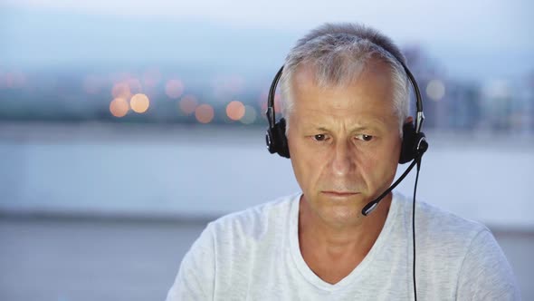 An Elderly Whitehaired Man Talks on the Internet on a Laptop with Headphones with a Microphone Late