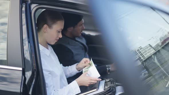 Side View Portrait of Slim Young Beautiful Woman Counting Cash Sitting in Car with Blurred Man alt
