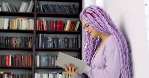 Young Woman with Long Colored Purple Hair is Reading Sitting in the Library
