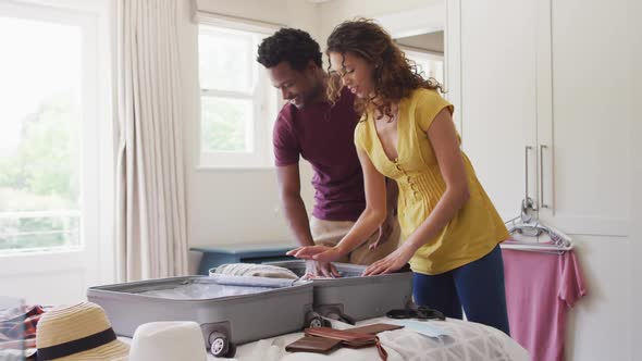 Happy biracial couple packing clothes into suitcase together, preparing for travel alt