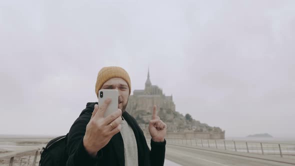 Happy Young Smiling Caucasian Blogger Man Making Video Call Using Smartphone at Mont Saint Michel alt