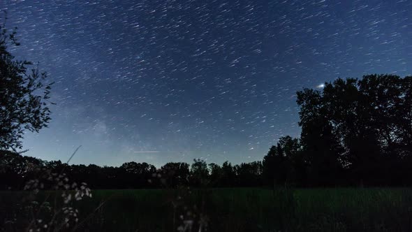 moving milky way and star trails on night sky over rural landscape with silhouette of trees alt