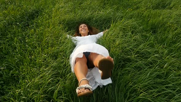 Cute Girl Falls Down Into the Green Grass. Happy Girl in White Dress Resting on Green Grass