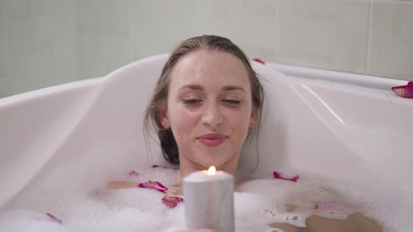 Portrait of Relaxed Smiling Beautiful Woman Lying in Water in Bathtub Blowing Out Candle in Slow alt