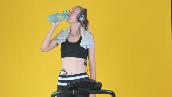 Fit blonde girl works out on a treadmill and drinking water from sport bottle.