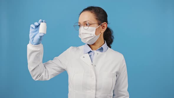Professional Woman Doctor Wearing Protective Mask Showing Jar with Pills at Camera Blue Studio alt