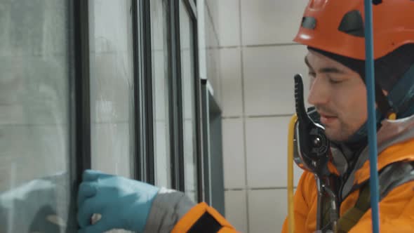 Window Cleanerin Helmet and Gloves Washes Windows with Rubber Screed Outside Apartment Building alt