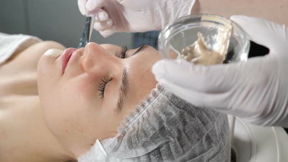 Cosmetician Applying Beauty Mask on Beautiful Female Girls Face. Cosmetic Golden Mask on Face alt