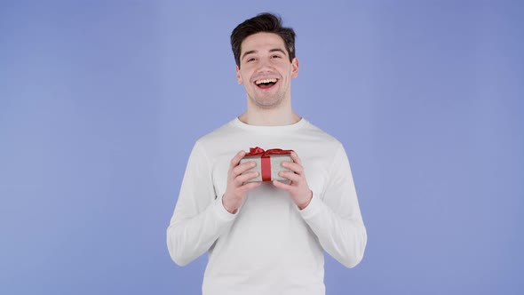 Handsome Man Holding Gift Box on Purple Studio Background and Smiles to Camera alt