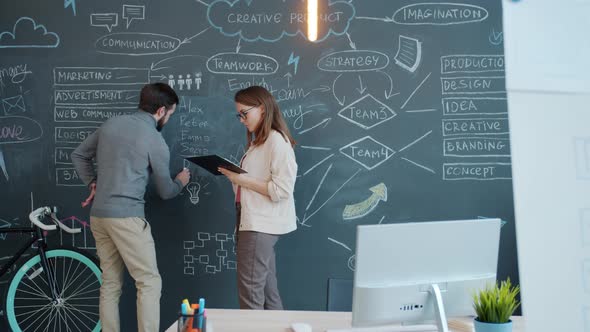Businesswoman Talking To Male Colleague Who Is Writing Information on Chalkboard in Office alt