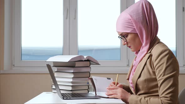 Muslim Woman Writes with a Pen in a Notebook Looking Into a Laptop Slow Motion alt