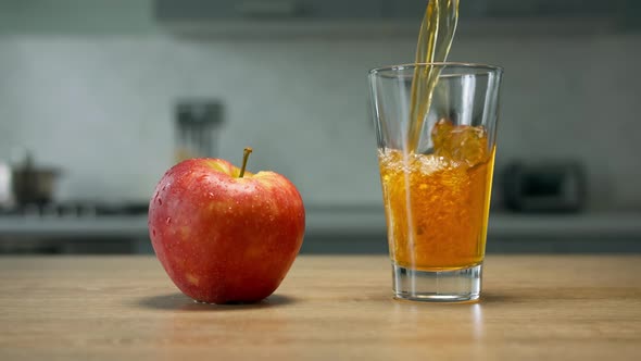 Apple juice is poured into glass on wooden table with red apple on it in the kitchen. Close up