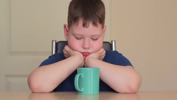 A Fat Child is Sitting at the Table Propping His Chin on His Hands Looking at a Mug