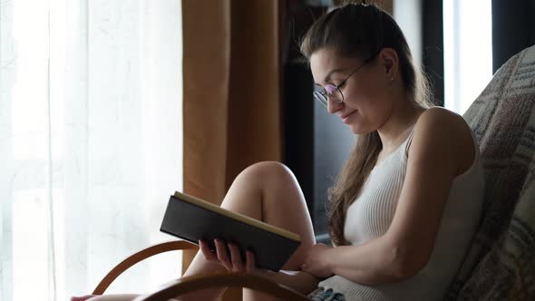 The girl in glasses reads a book in natural light near the window and chuckles alt