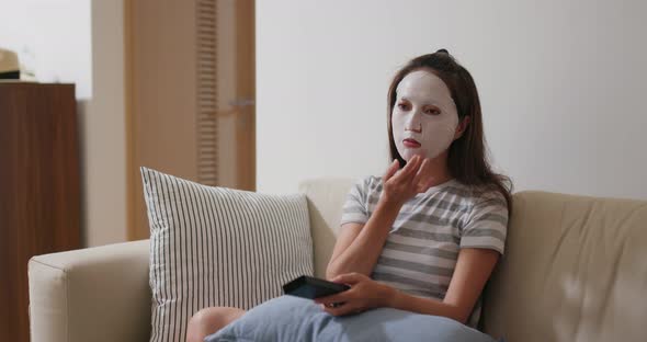 Woman apply face mask and watch TV at home alt