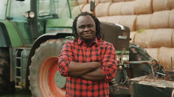 Young African Farmer Nodding Head As Approval with Crossed Hands Over the Chest in Front of Tractor alt