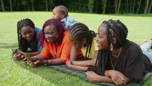 Cheerful Black Family Lying on Plaid and Sharing Tablet Pc in Park alt