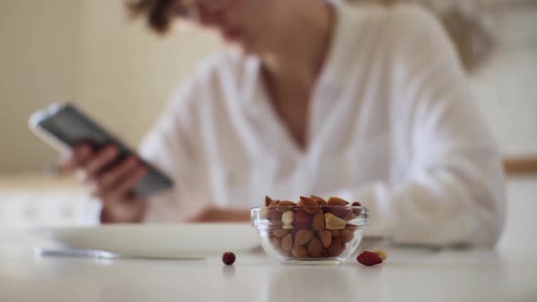 Bowl Of Mixed Nuts, In Foreground On Desktop. Healthy Food And Work. Woman Eats Nuts While Working alt