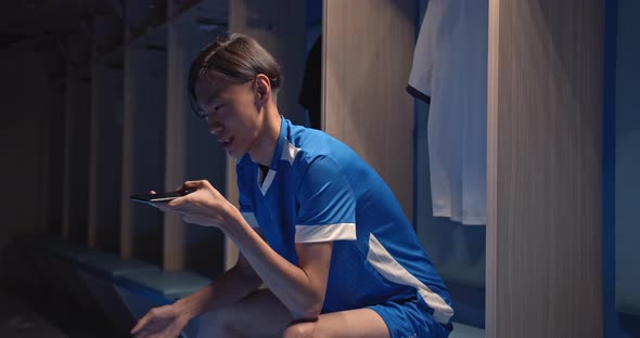 Asian Young Soccer Player Man Talking with Phone Sitting in the Locker Room at the Gym or Stadium