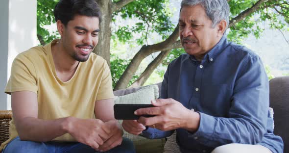 Man sharing smart phone with young son while sitting together at patio alt