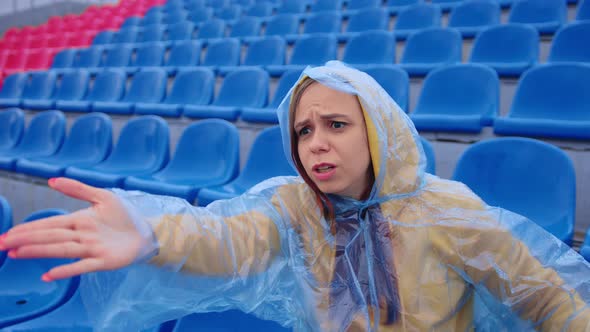 Unhappy Woman in Raincoat Sitting on Stadium Bleachers Alone Rooting for Favorite Sports Team alt