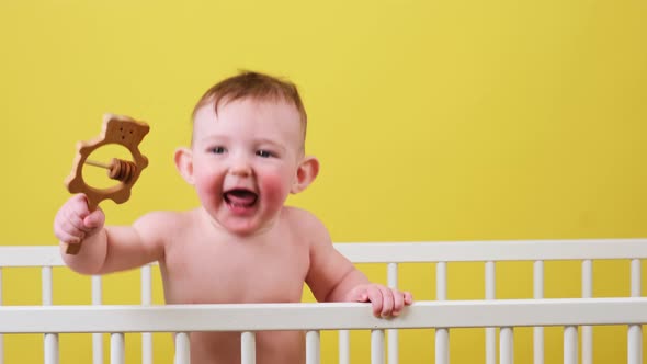 Happy toddler baby boy in a crib with a wooden toy in his hand, yellow studio background. A smiling