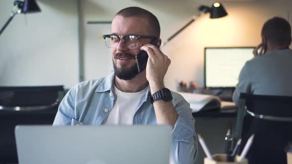 Young Businessman is Talking on Phone at Table with Laptop in White Office alt
