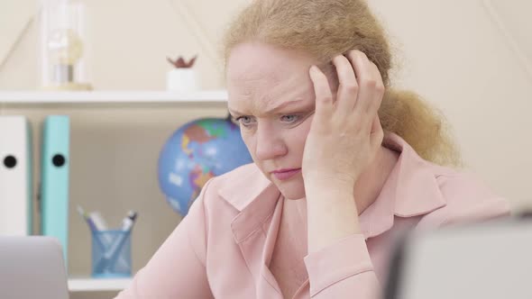 Close-up Face of Desperate Caucasian Senior Woman Holding Head with Hand As Looking at Laptop Screen alt