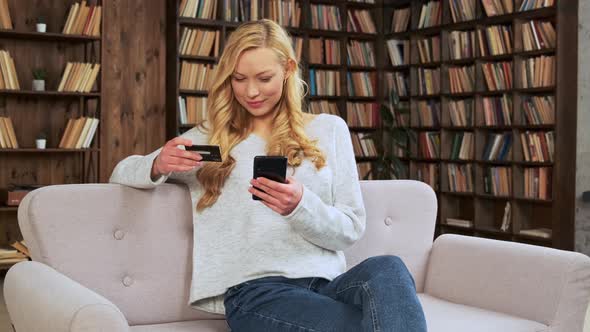 Young Blonde Woman Sitting on the Couch and Makes Purchases Online Using Credit Card and Smart Phone alt
